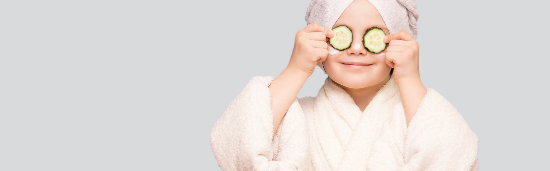 Funny little girl in white coat and towel with mask for skin face and cucumbers on eyes isolated on white background. Spa skin care treatments